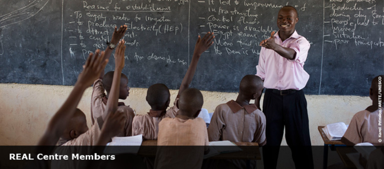 teacher in classroom in from of chalk board with children holding their hands up to answer a question