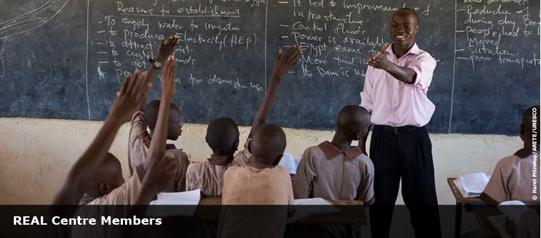 teacher in classroom in from of chalk board with children holding their hands up to answer a question