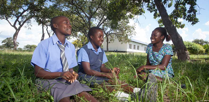 two school pupils and a teacher sit under a tree with a school building in the background